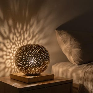 Könnte beinhalten: Dekorative, kugelförmige Lampe mit perforiertem Metallschirm, die ein gemustertes Licht an die Wand wirft. Die Lampe steht auf einem quadratischen Holzsockel, der auf einem dunklen Nachttisch aus Holz platziert ist. Das warme Licht erzeugt ein gemütliches Ambiente.