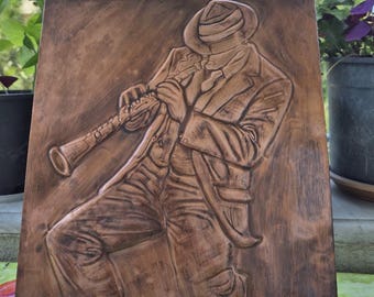 May include: A rectangular wooden plaque featuring a relief carving of a musician wearing a fedora and a suit, playing a clarinet. The artwork is finished in a medium brown stain with darker shading to highlight the carved details of the figure.