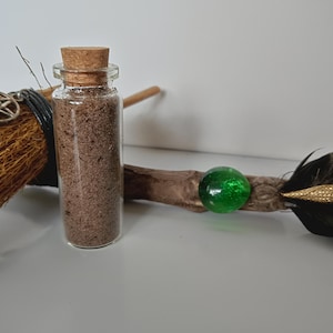 May include: A small glass bottle with a cork stopper filled with brown granules. A decorative broom and a wand with a green glass orb and black feathers are in the background. The items are on a white surface.