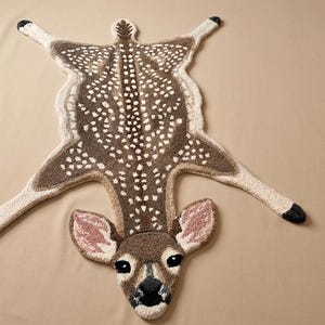 Può includere: Un tappeto tufted a forma di cerbiatto, caratterizzato da un corpo marrone con macchie bianche, una striscia dorsale marrone scuro e quattro zampe con zoccoli neri. La testa presenta grandi orecchie con dettagli rosa, occhi e naso neri.