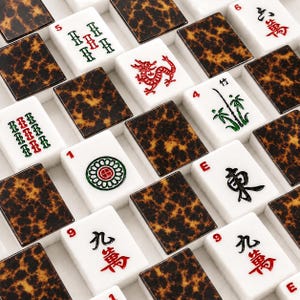 May include: A collection of Mahjong tiles arranged in a grid pattern. The tiles feature white faces with various red, green, and black symbols, numbers, and characters, alternating with dark brown and black tortoiseshell patterned backs.
