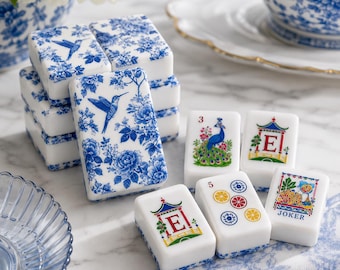 May include: A set of white mahjong tiles featuring blue floral and hummingbird patterns, alongside tiles decorated with colorful illustrations of a peacock, a pagoda with the letter E, citrus slices, and a joker card with a floral design.