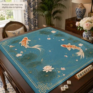 Puede incluir: Un mantel cuadrado de color verde azulado con dos peces koi naranjas y blancos nadando entre nen&uacute;fares de color verde oscuro y flores de loto blancas. El dise&ntilde;o incluye l&iacute;neas doradas y un borde geom&eacute;trico. El color puede variar ligeramente.