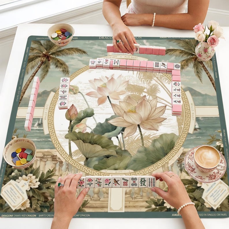 May include: A square mahjong mat featuring a central illustration of lotus flowers and palm trees in muted green and cream tones. The mat includes game-specific text, borders, and a circular frame. Pink mahjong tiles are arranged in rows.