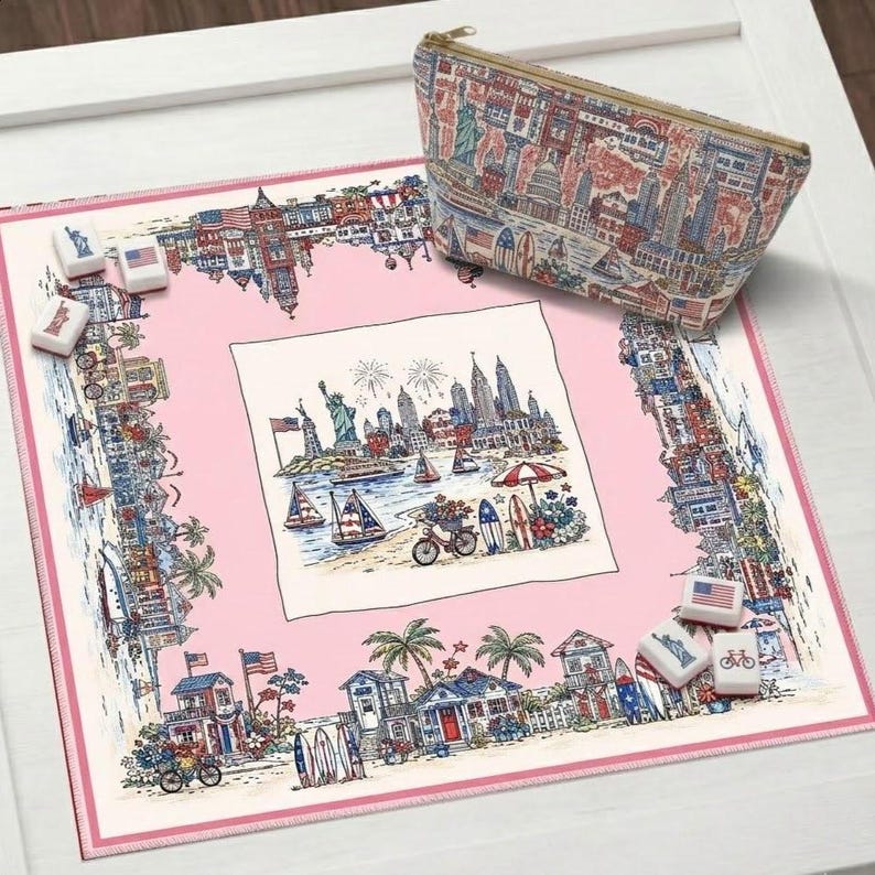 May include: A square fabric mat with a pink border and a central illustration of a coastal city skyline with the Statue of Liberty, sailboats, and a bicycle. A matching zippered pouch features a repeating city pattern, alongside small white tiles.
