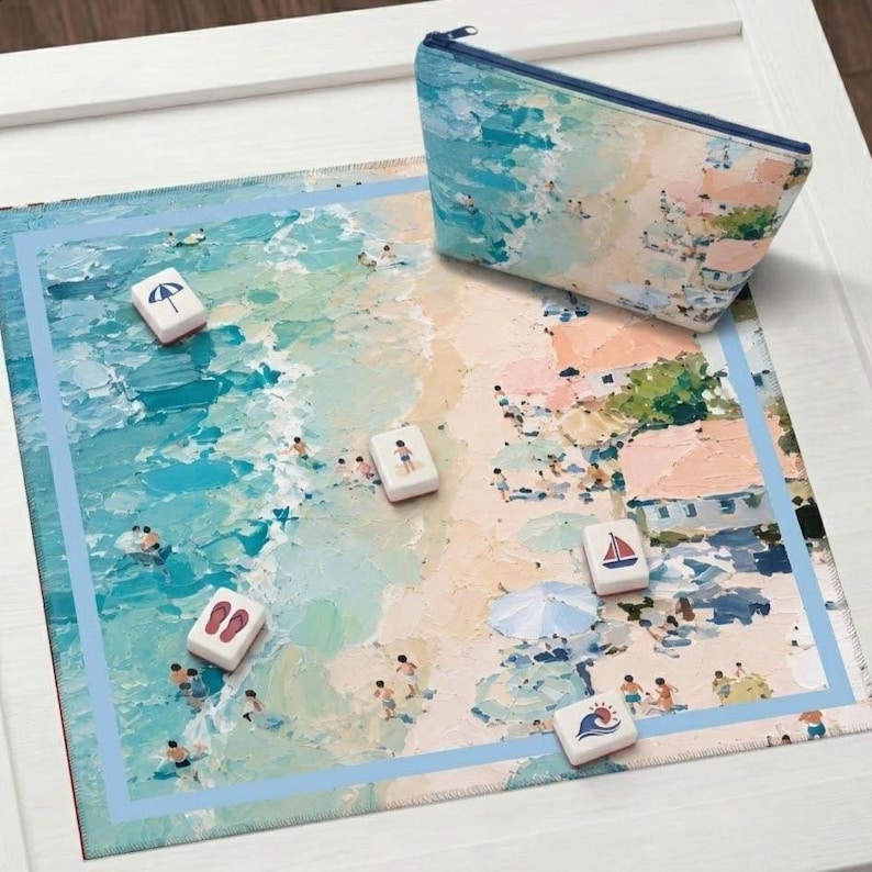 May include: A beach-themed mat and matching zipped pouch. The mat depicts a beach scene with people, water, and sand. Several white game tiles with beach-related icons are scattered on the mat. The pouch has a similar beach scene design.