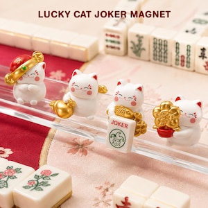 May include: Four white lucky cat figurines arranged in a row. The first wears a red hat, the second holds a gold bag, the third holds a gold fish and a white mahjong tile labeled JOKER, and the fourth holds a gold flower. Text reads LUCKY CAT JOKER MAGNET.