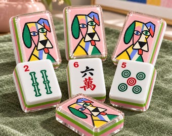 May include: A set of seven mahjong tiles featuring three tiles with a colorful, abstract dog illustration on a pink background, and four tiles with traditional green and red symbols, including the numbers 2, 5, and 6 in black and red ink.