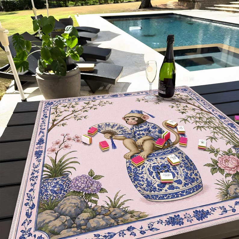 May include: A pink and blue decorative table mat featuring a monkey in a blue and white outfit, holding a fan and surrounded by game tiles. The mat has a floral and botanical design with a blue border.