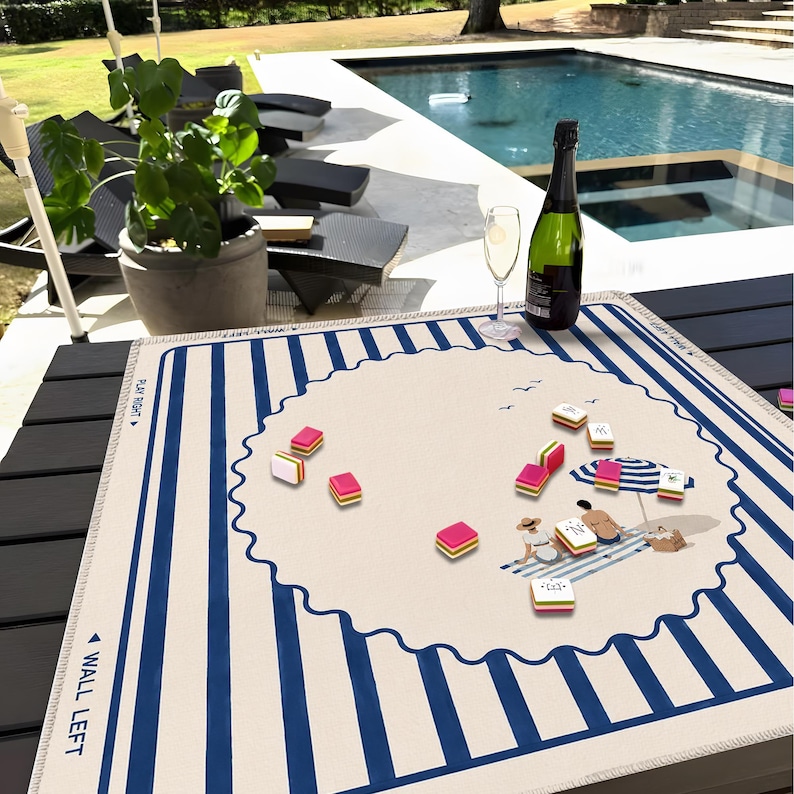 May include: A Mahjong game set on a striped blue and white mat with a beach scene illustration. The set includes pink and yellow tiles, a bottle of champagne, and a glass. The mat has the words "WALL LEFT" and "PLAY RIGHT".