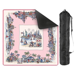 May include: A square picnic blanket with a light pink border and a central illustration of the Statue of Liberty, city skyline, sailboats, and beach scenes. The blanket has a black underside and comes with a black cylindrical carrying bag.