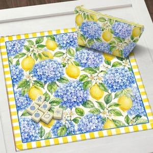 May include: A floral pattern featuring blue hydrangeas and yellow lemons adorns a rectangular mat and a zippered pouch. The mat has a yellow and white striped border. Several mahjong tiles are visible on the mat.