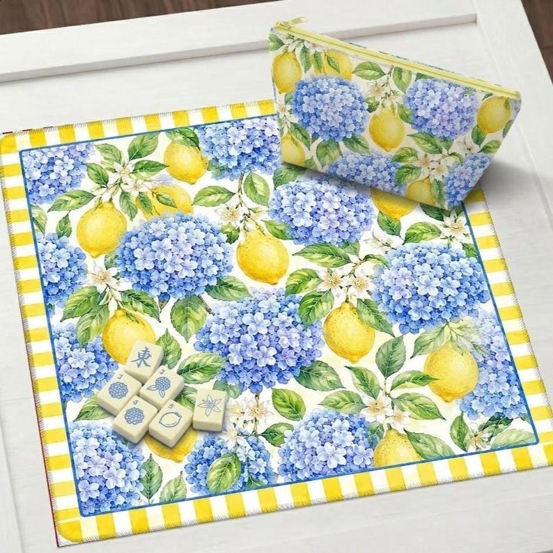 May include: A floral pattern featuring blue hydrangeas and yellow lemons adorns a rectangular mat and a zippered pouch. The mat has a yellow and white striped border. Several mahjong tiles are visible on the mat.