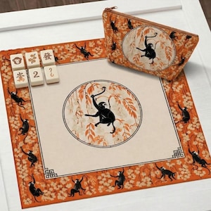 Op de afbeelding: Een decoratieve set met een apen thema. Inclusief een rechthoekige mat met een centraal cirkelvormig ontwerp en een bijpassend etui met ritssluiting. De mat en het etui hebben een oranje rand met apen- en bloempatronen. Er zijn ook verschillende Mahjong-tegels aanwezig.