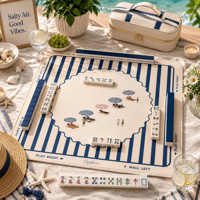 May include: A mahjong set with a beach theme. The game board is cream-colored with blue stripes and a scalloped edge. The center features a beach scene with umbrellas and people. The set includes tiles, racks, and a matching travel case.