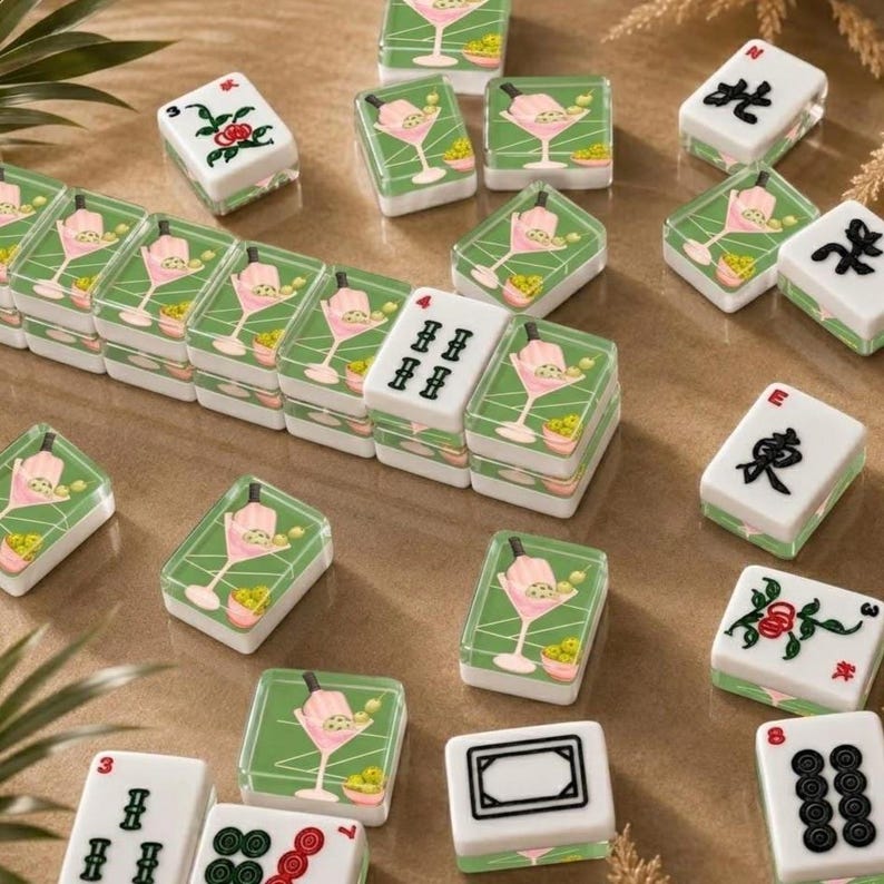 May include: A collection of decorative Mahjong tiles. The tiles feature a green background with a pink cocktail design and olives. Some tiles have black symbols and numbers. The tiles are arranged on a light brown surface.