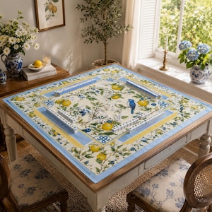 Puede incluir: Una mesa cuadrada de madera con un mantel floral con limones, ar&aacute;ndanos y un p&aacute;jaro azul. La mesa est&aacute; preparada para jugar al Mahjong, con fichas y soportes visibles. Las sillas tienen tapicer&iacute;a floral a juego.