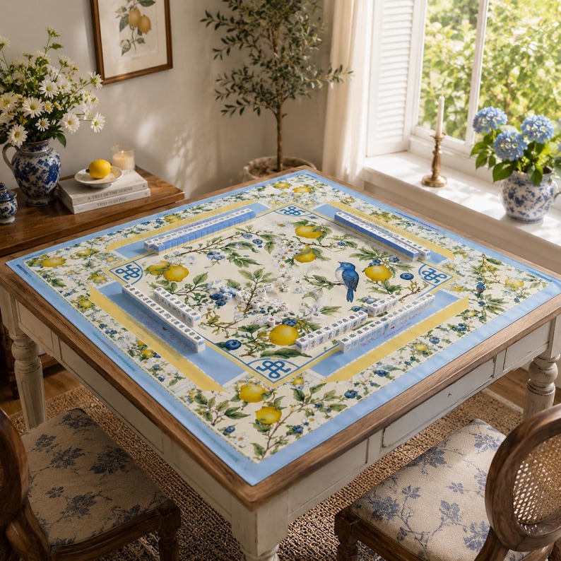 Puede incluir: Una mesa cuadrada de madera con un mantel floral con limones, ar&aacute;ndanos y un p&aacute;jaro azul. La mesa est&aacute; preparada para jugar al Mahjong, con fichas y soportes visibles. Las sillas tienen tapicer&iacute;a floral a juego.