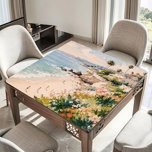 May include: A square wooden table with a painted beach scene on the tabletop. The artwork depicts a sandy beach, ocean waves, and palm trees. The table has decorative cutouts on the sides and is surrounded by neutral-colored chairs.