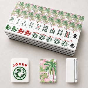 May include: A complete Mahjong set with white tiles featuring green and red dragon designs, numbers, and Chinese characters. The set includes a box with a pink and green palm tree pattern. Additional tiles and a lighter are also visible.