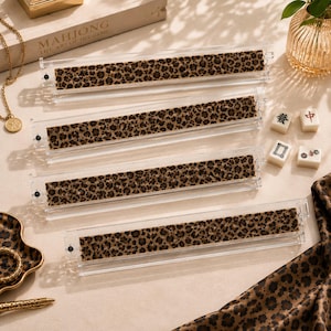 May include: Clear acrylic Mahjong tile racks with a leopard print design. The rectangular racks hold tiles for the game. The image also shows Mahjong tiles and a box with the words "Mahjong The Art of the Game".