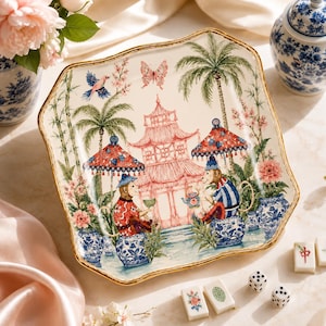 Könnte beinhalten: Ein quadratischer Keramikteller mit gewelltem Rand und Goldkante, der eine detaillierte Illustration von zwei Affen zeigt, die auf blau-weißen Hockern unter Sonnenschirmen sitzen, mit einer rosa Pagode, Palmen, Blumen und einem Schmetterling.