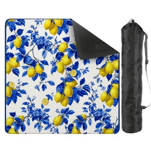 May include: A picnic blanket with a white background, featuring a pattern of yellow lemons and blue leaves and flowers. The blanket has a black underside and comes with a black carrying bag.