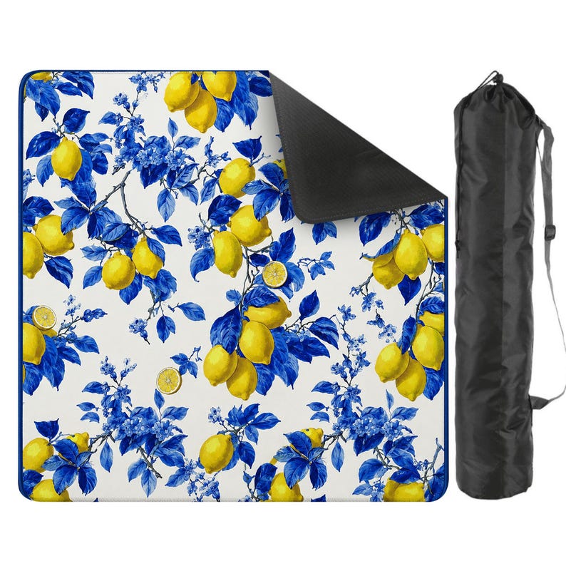 May include: A picnic blanket with a white background, featuring a pattern of yellow lemons and blue leaves and flowers. The blanket has a black underside and comes with a black carrying bag.