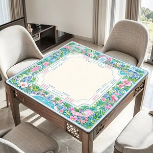 May include: A square mahjong mat featuring a cream-colored center with printed game instructions and a vibrant border of pink hibiscus flowers, green palm leaves, blue and white porcelain vases, pink flamingos, and striped lounge chairs.