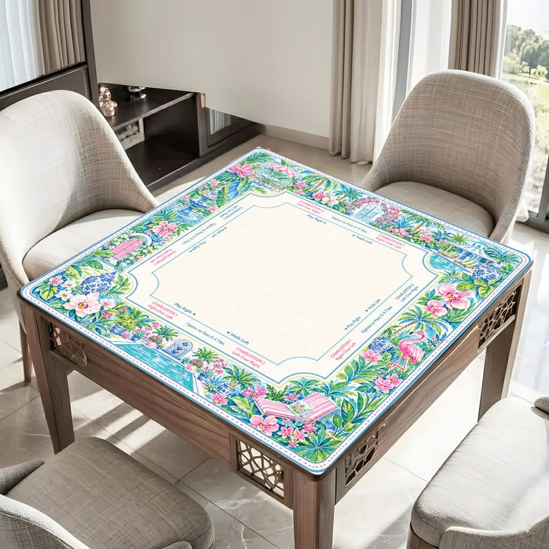 May include: A square mahjong mat featuring a cream-colored center with printed game instructions and a vibrant border of pink hibiscus flowers, green palm leaves, blue and white porcelain vases, pink flamingos, and striped lounge chairs.
