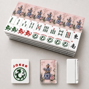 Op de afbeelding: Een complete Mahjong-set met witte tegels met zwarte symbolen en rode of groene drakenontwerpen. De set bevat een roze koffer met een decoratief patroon en accessoires, waaronder een Joker-tegel en een decoratieve kaart.