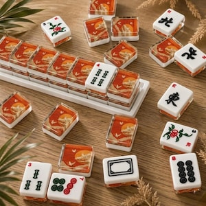 May include: A set of Mahjong tiles with a sunset design. The tiles are white with orange and clear accents. Some tiles have Chinese characters, while others have flower and dot designs. The tiles are arranged on a wooden surface.