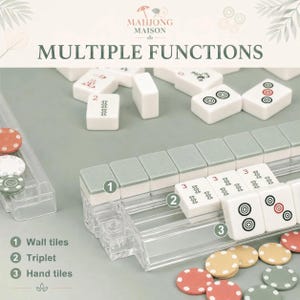 May include: A Mahjong Maison set featuring white rectangular tiles with black and red symbols, sage green tiles, and small circular tokens in red, white, and green. A clear plastic rack holds a row of tiles, with text labels for wall tiles, triplet, and hand tiles.