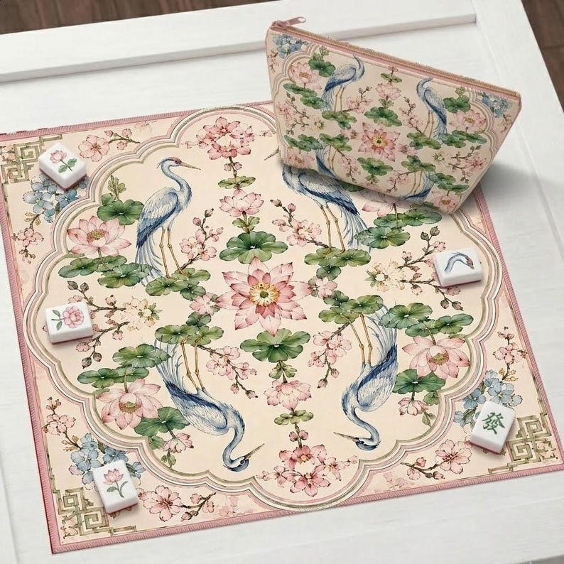 May include: A decorative set featuring a floral and bird design. The set includes a patterned mat, a matching zippered pouch, and several Mahjong tiles. The design features blue herons, pink flowers, and green leaves on a cream background.
