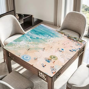 May include: Square wooden table featuring a beach scene print. The artwork shows a sandy beach, turquoise water, and people with parasols. The table has decorative cutouts on the sides. Chairs surround the table.