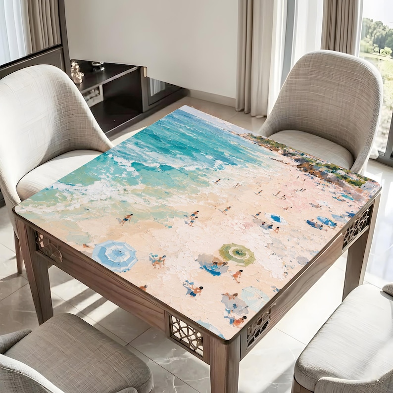 May include: Square wooden table featuring a beach scene print. The artwork shows a sandy beach, turquoise water, and people with parasols. The table has decorative cutouts on the sides. Chairs surround the table.