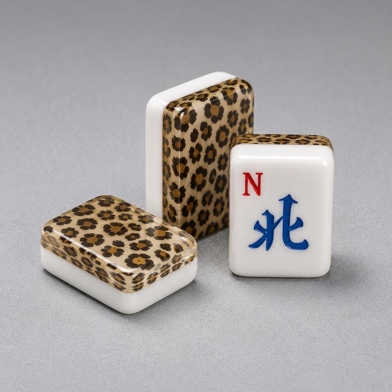 Puede incluir: Tres fichas de Mahjong. Dos fichas tienen un dise&ntilde;o de estampado de leopardo en la superficie superior. La tercera ficha es blanca con un s&iacute;mbolo azul y rojo y la letra "N". Las fichas est&aacute;n sobre una superficie gris.