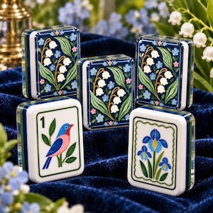 Puede incluir: Cinco fichas de mahjong rectangulares con bordes redondeados. Tres fichas presentan fondos azul oscuro con lirios de los valles blancos y pequeñas flores azules. Una ficha muestra un pájaro azul y rosa con el número 1. Una ficha muestra un iris azul.