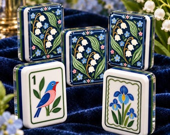 May include: Five rectangular mahjong tiles with rounded edges. Three tiles feature dark blue backgrounds with white lily of the valley and small blue flowers. One tile displays a blue and pink bird with the number 1. One tile shows a blue iris.