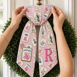 May include: A pink fabric bow featuring a repeating pattern of Mahjong tiles, including Joker cards with pink monkeys, peacocks, and floral designs. The right side of the bow displays a large, embroidered pink letter R in a serif font.