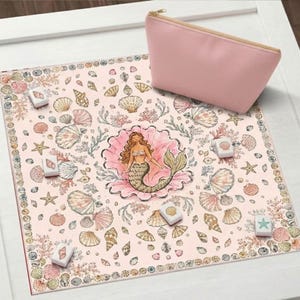 May include: A square light pink fabric mat featuring a central illustration of a mermaid inside a shell, surrounded by various seashells, starfish, and coral patterns. Five small white square tiles with shell and starfish icons are placed on the mat, accompanied by a matching light pink zippered pouch.