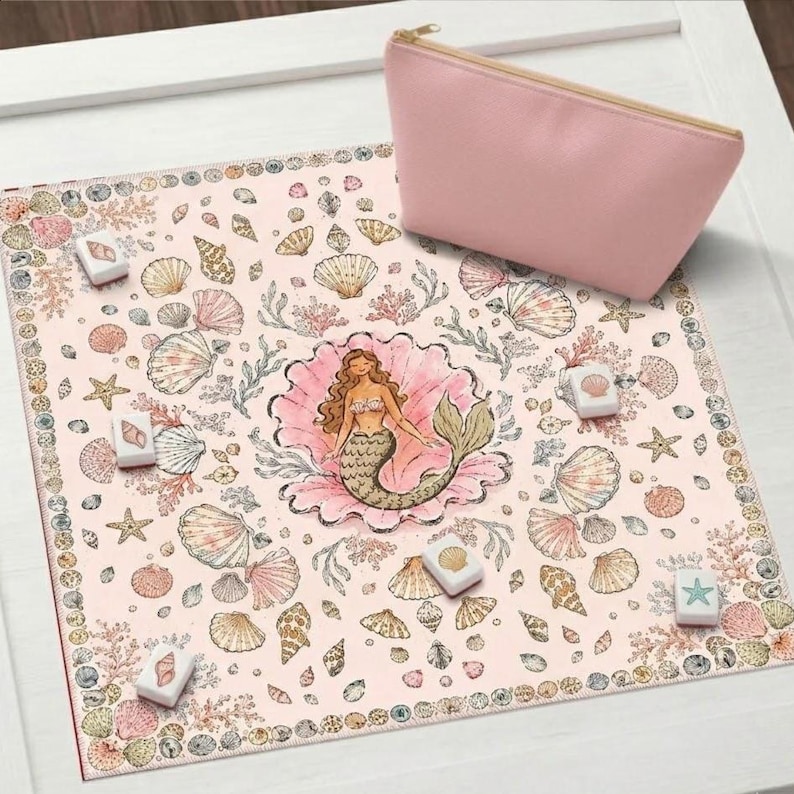May include: A square light pink fabric mat featuring a central illustration of a mermaid inside a shell, surrounded by various seashells, starfish, and coral patterns. Five small white square tiles with shell and starfish icons are placed on the mat, accompanied by a matching light pink zippered pouch.