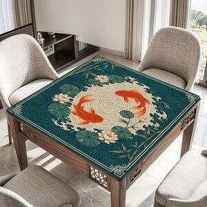 May include: A square table mat featuring a dark teal background with a central cream-colored circle. Two orange koi fish swim in a circular pattern surrounded by white lotus flowers, green lily pads, bamboo stalks, and stylized wave motifs.