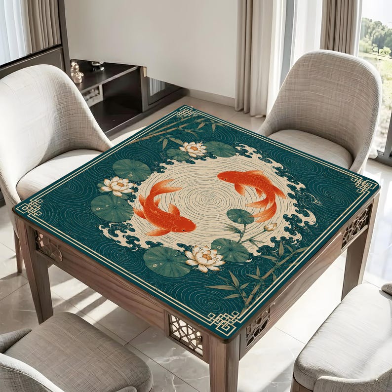 May include: A square table mat featuring a dark teal background with a central cream-colored circle. Two orange koi fish swim in a circular pattern surrounded by white lotus flowers, green lily pads, bamboo stalks, and stylized wave motifs.