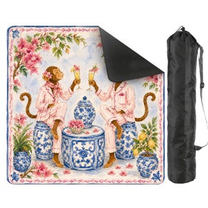 May include: A picnic blanket featuring a whimsical illustration of two monkeys in pink pajamas toasting cocktails. The blanket has a blue floral border and comes with a black carrying bag. The design includes blue and white ceramic pots and pink flowers.