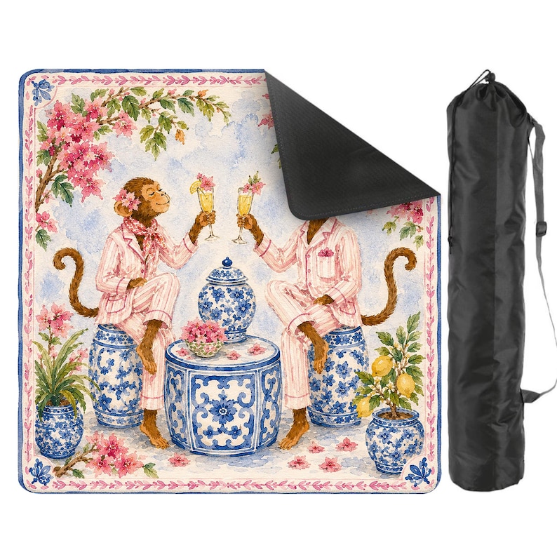 May include: A picnic blanket featuring a whimsical illustration of two monkeys in pink pajamas toasting cocktails. The blanket has a blue floral border and comes with a black carrying bag. The design includes blue and white ceramic pots and pink flowers.
