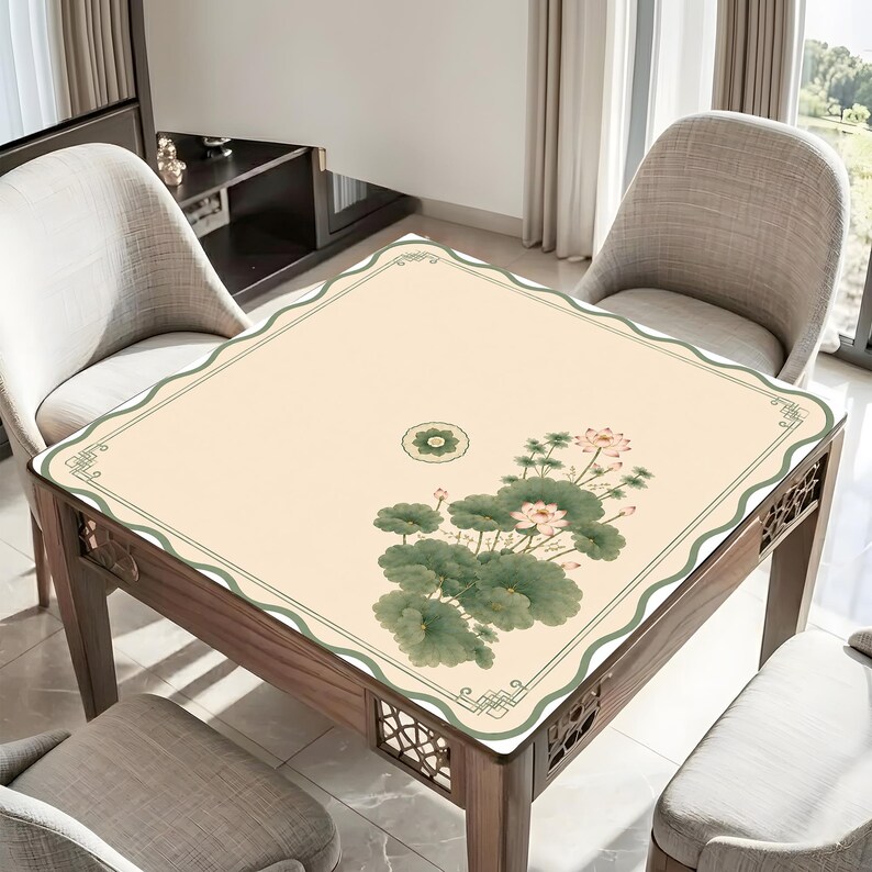May include: A square table mat with a cream-colored surface featuring a green wavy border and a decorative corner pattern. The mat displays a printed illustration of green lotus leaves and pink lotus flowers, along with a small circular floral motif.