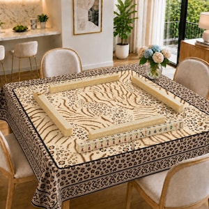 May include: A square mahjong table covered with a beige tablecloth featuring a brown leopard and tiger stripe pattern. Four wooden racks are arranged in a square on the table, with one rack displaying a row of mahjong tiles with red and black symbols.