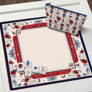 Op de afbeelding: Een vierkante speelmat met een crèmekleurig midden en een rand met rode rozen, witte bloemen, blauwe vazen en Amerikaanse vlaggen, vergezeld door een bijpassend tasje met rits en vijf witte rechthoekige tegels met bedrukte ontwerpen.