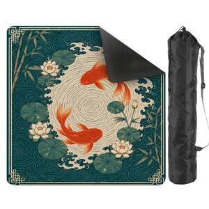 May include: A square outdoor mat featuring a dark teal background with a pattern of two orange koi fish, white lotus flowers, and green lily pads. The mat has a cream-colored circular center with ripple lines and a black carrying bag with a shoulder strap.